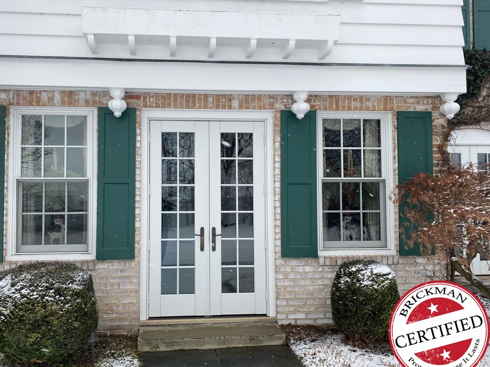 The French door entrance of the Hickory Corners house in 2021, white brick between green shutters with snow on the boxwoods and the certification stamp in the corner