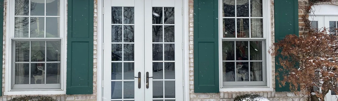 The French door entrance of the Hickory Corners house in 2021, white brick between green shutters with light snow on the boxwoods