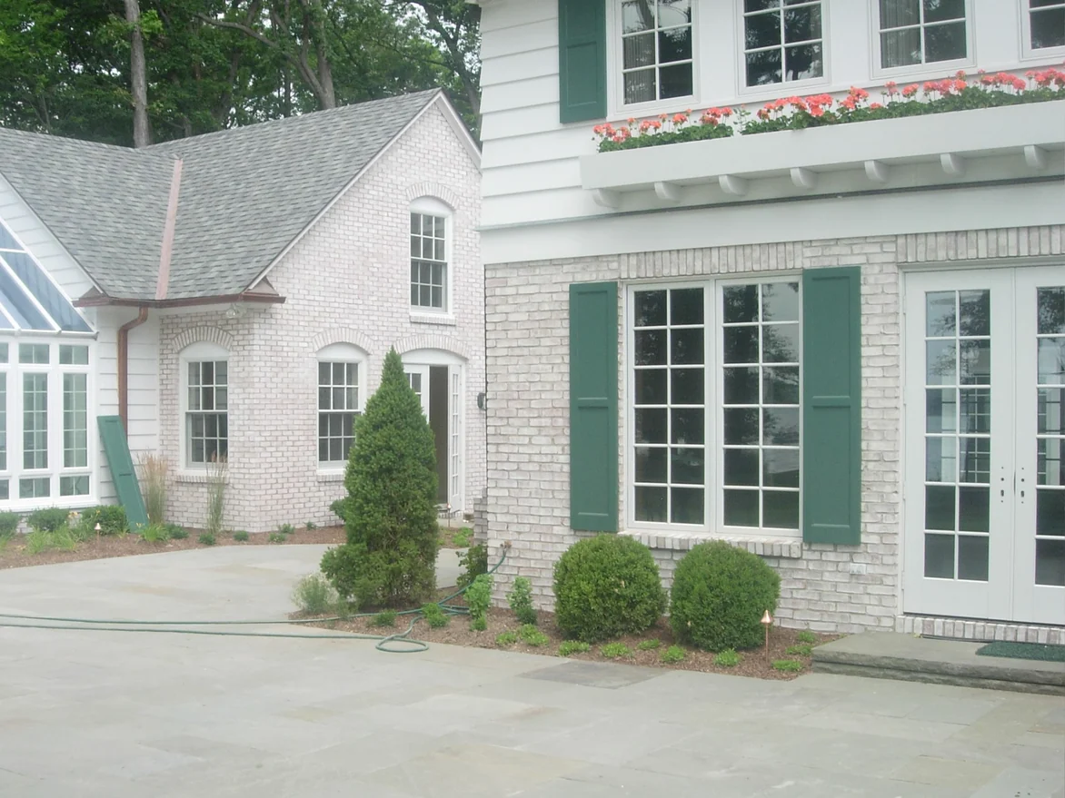 The same shuttered window after staining, the brick now white, with a gabled brick wing and a glass conservatory beyond it across a stone patio