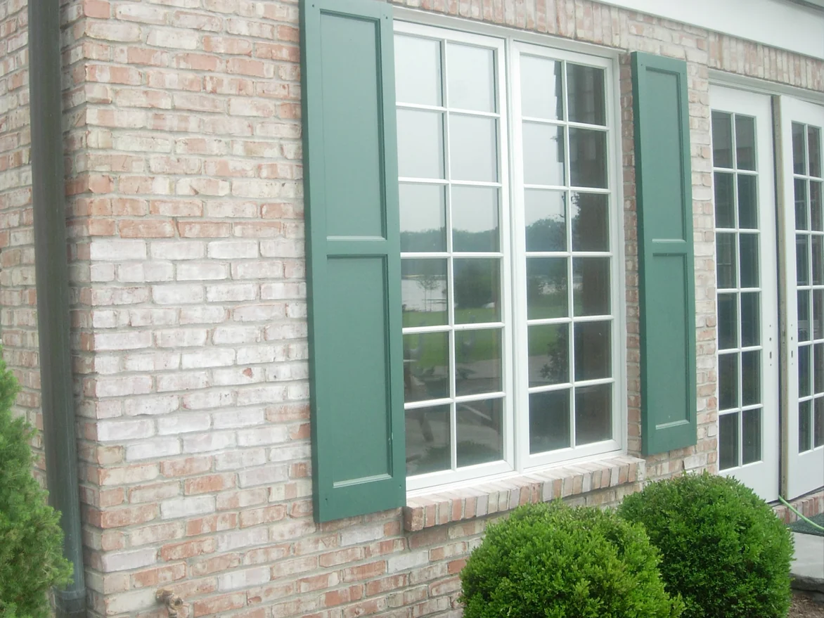 A corner of the house before staining, tumbled pink and red brick with pale flecks around a divided-light window between green shutters, a downspout at the corner and boxwoods below