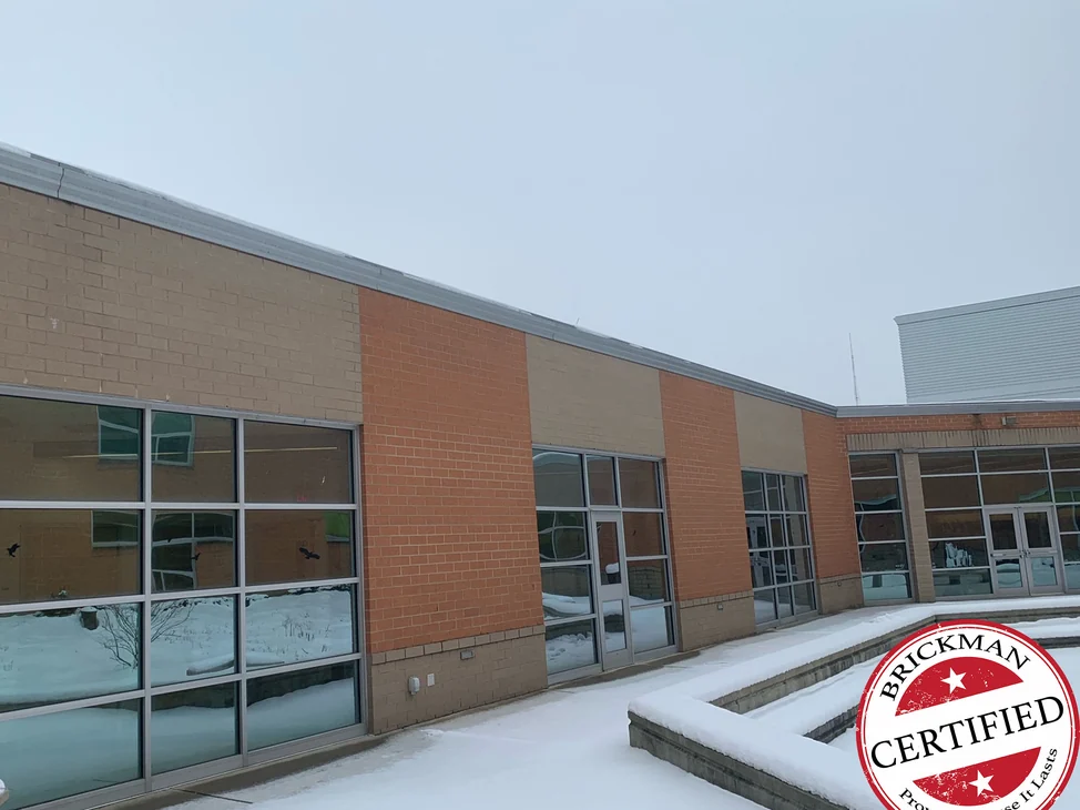 The high school courtyard in 2021 under snow, tan panels above each window bay between piers of orange brick, with the certification stamp in the corner.