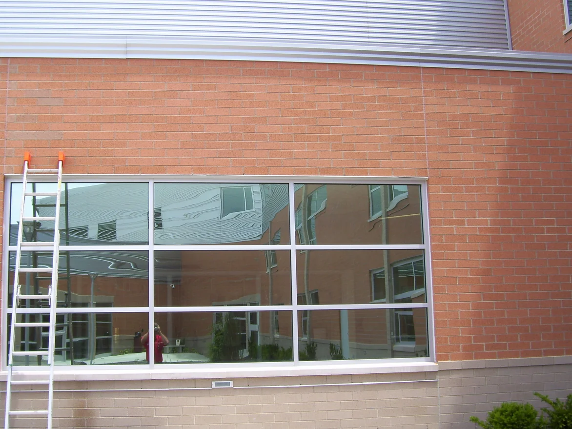 A courtyard window bay in 2011 before staining, the wall above and beside it laid entirely in orange brick under a corrugated metal parapet, with an extension ladder leaning on the glass