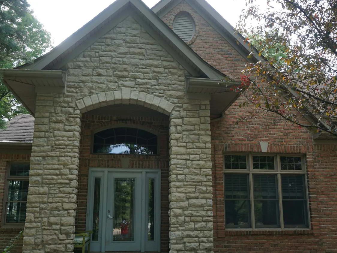 The front entry of the house in 2019, a gabled porch of split-face stone with an arched opening and a glazed door, red brick walls and a three-part window to the right