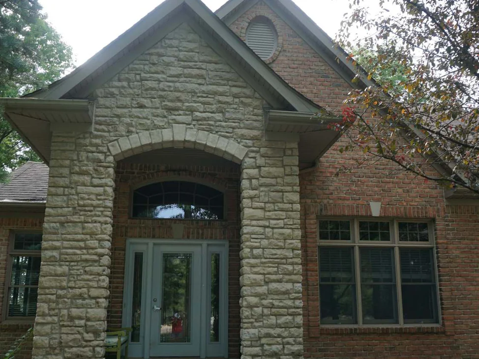 The front entry of the Highland house in 2019, a gabled porch of split-face stone with an arched opening, red brick walls and a three-part window to the right