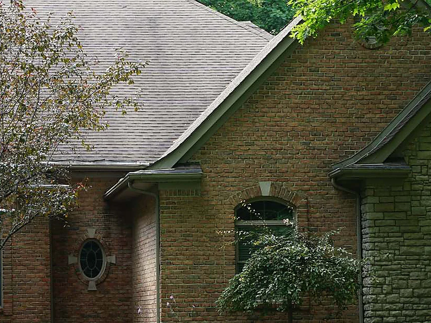 The front gable in 2019 seen through summer trees, the arched window and the oval window in one range of red brick beside the stone entry