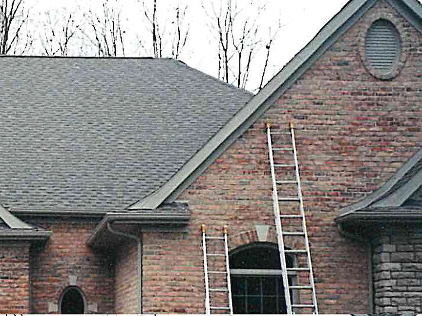 The front gable of the house in 2000 with two extension ladders leaning against the red brick beside an arched window, bare trees behind the roof