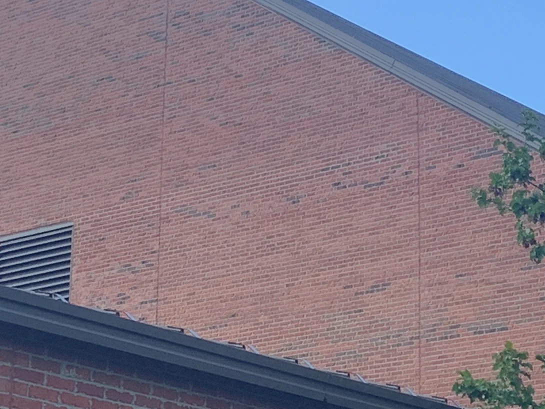 The upper gable wall in 2019, scattered darker header bricks reading through an even orange field under the eaves