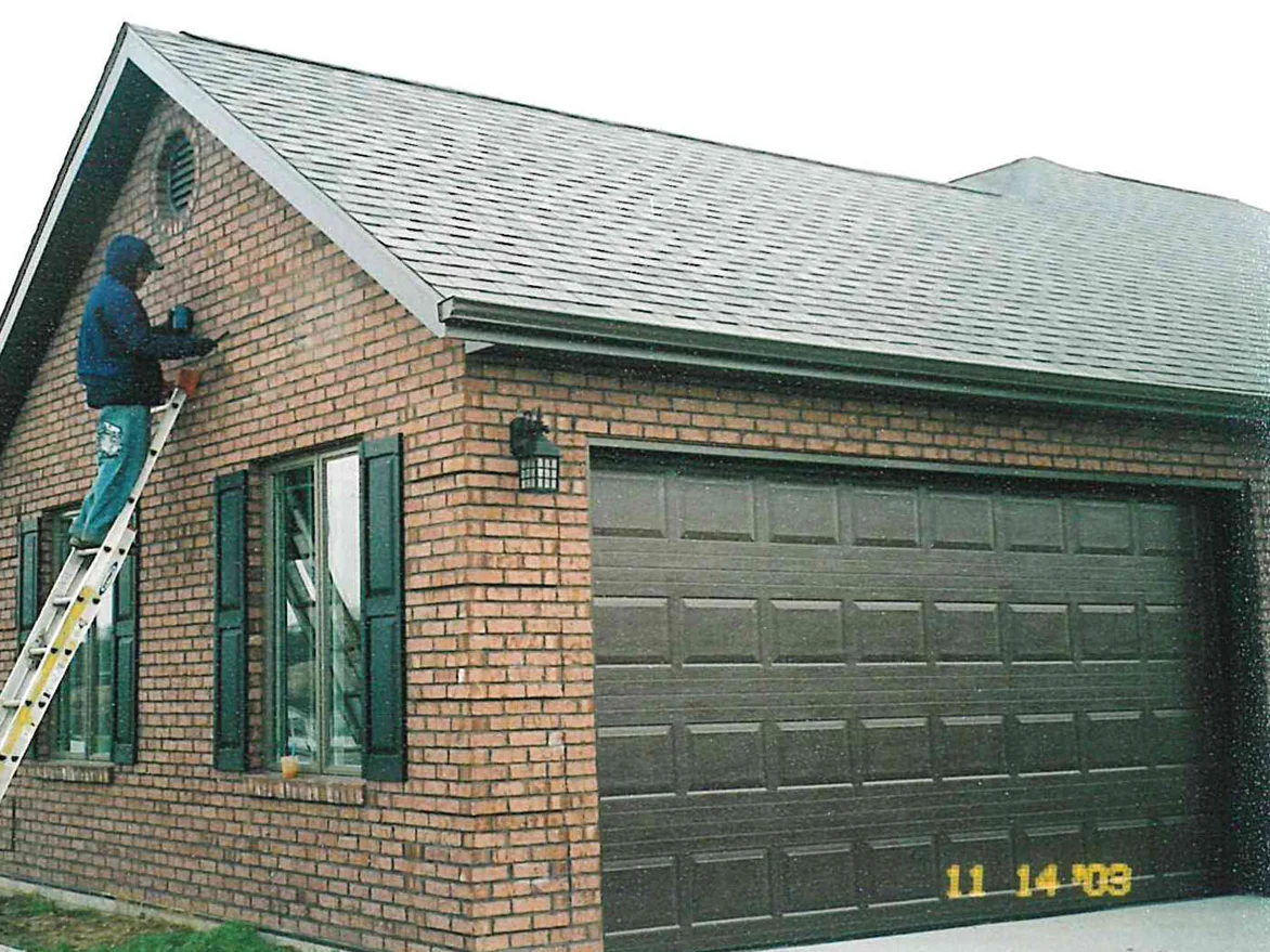 An applicator on a ladder working across the gable beside the garage door, the brick a mottled orange and tan with dark units through it