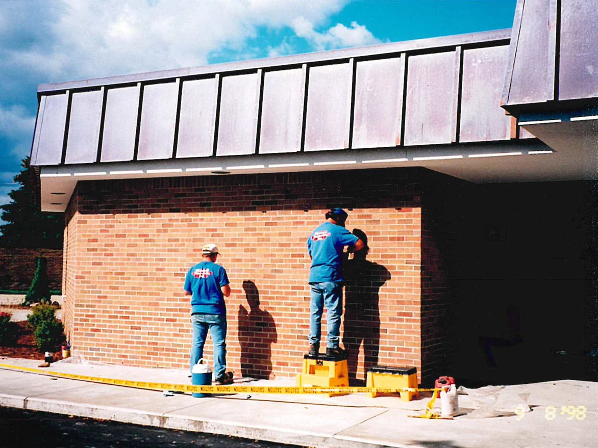 Two applicators working stain into the new brick of the addition, one from the pavement and one from a step stool, the ground taped off below them