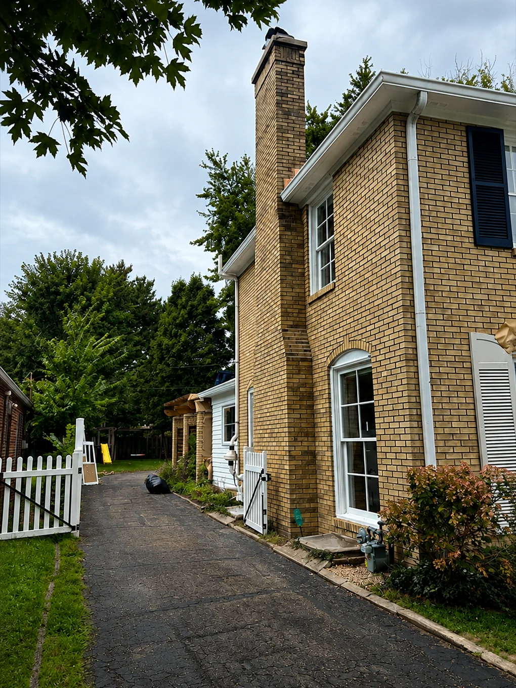The side of the house from the driveway, buff brick with the chimney running the full height of the gable
