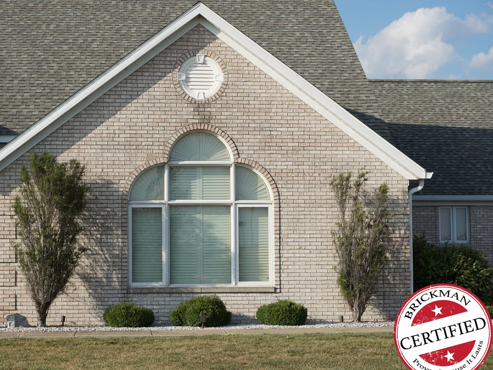The front gable of the house in 2019, its arched window and round louvered vent set in brick that reads as one cream and grey range, with the certification stamp at the corner of the frame.