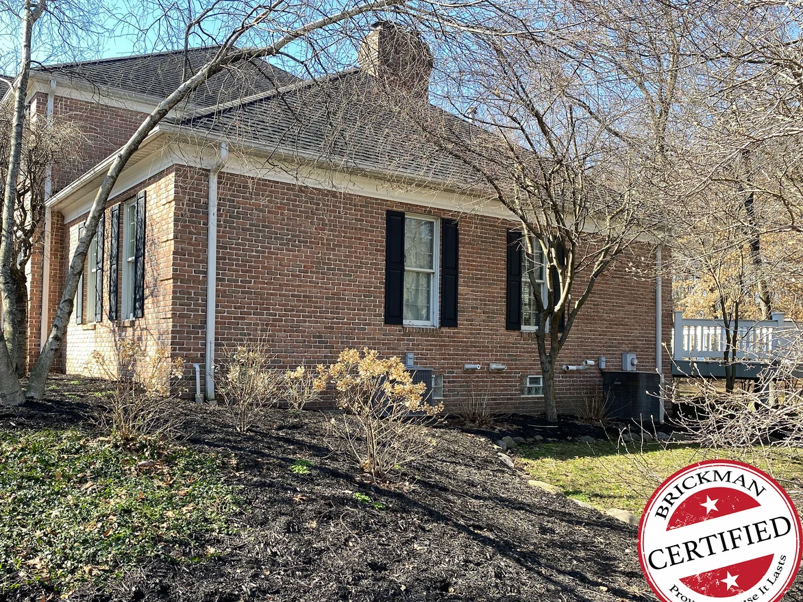 The side of the house in 2021, red brown brick running past shuttered windows to a chimney above the hipped roof, with the certification stamp in the corner