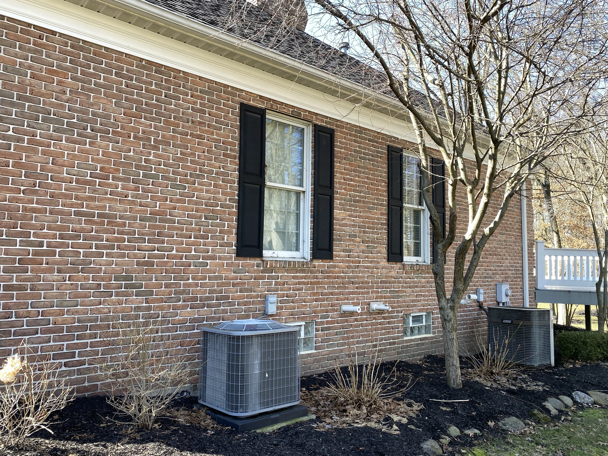 The side wall in 2021, shuttered windows over two outdoor condensing units, the brick reading as one range from the eaves down to the basement course