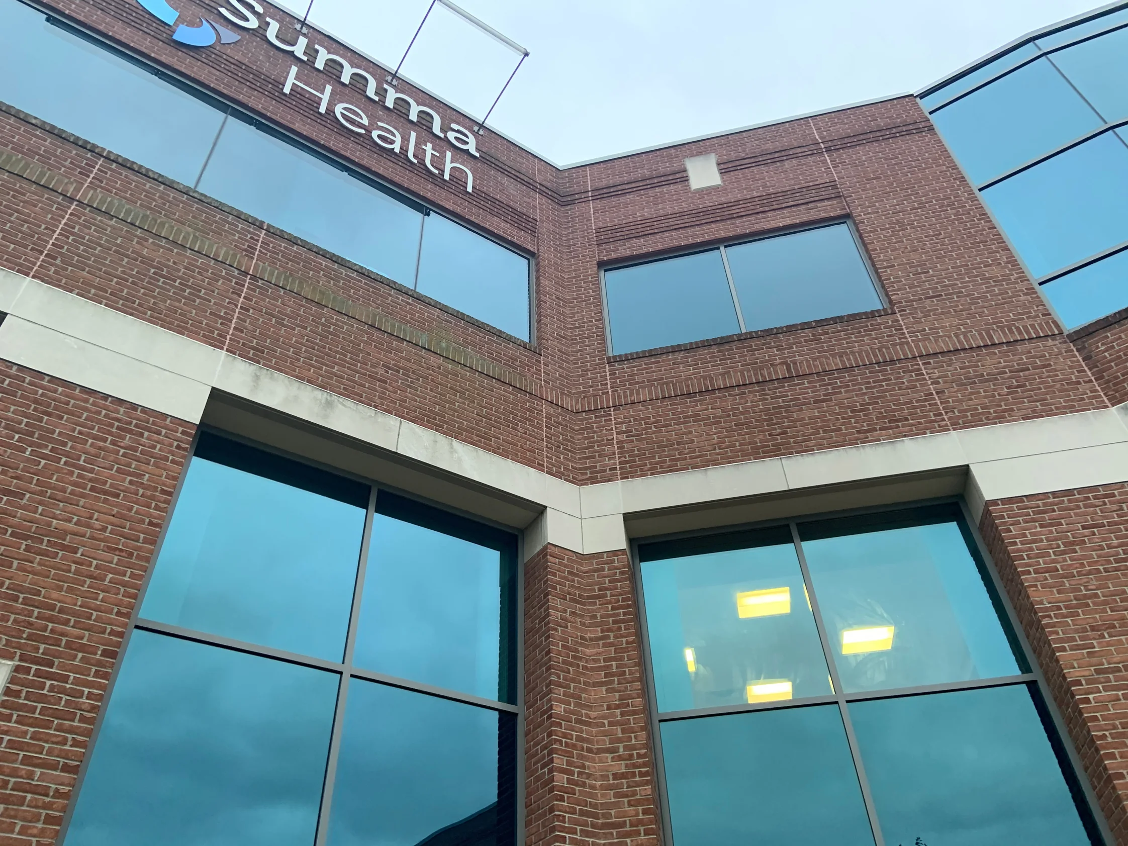 Looking up at the clipped corner of the building in 2019, red brick banding above two upper windows, a cast stone band over the ground floor glazing and a Summa Health sign on the parapet