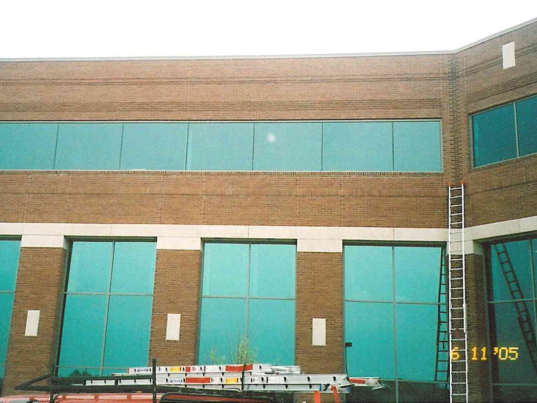 The upper elevation of the building in June 2005, tan brick with projecting bands above a ribbon of green glass, ladders laid across a van roof below and one extension ladder set against the corner