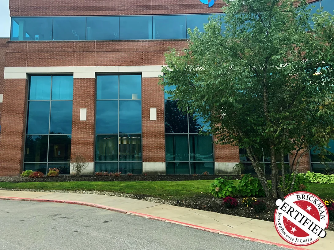 The front of the building in 2019, two tall window bays between red brick piers under a glazed upper floor, a tree in front and the certification stamp in the corner