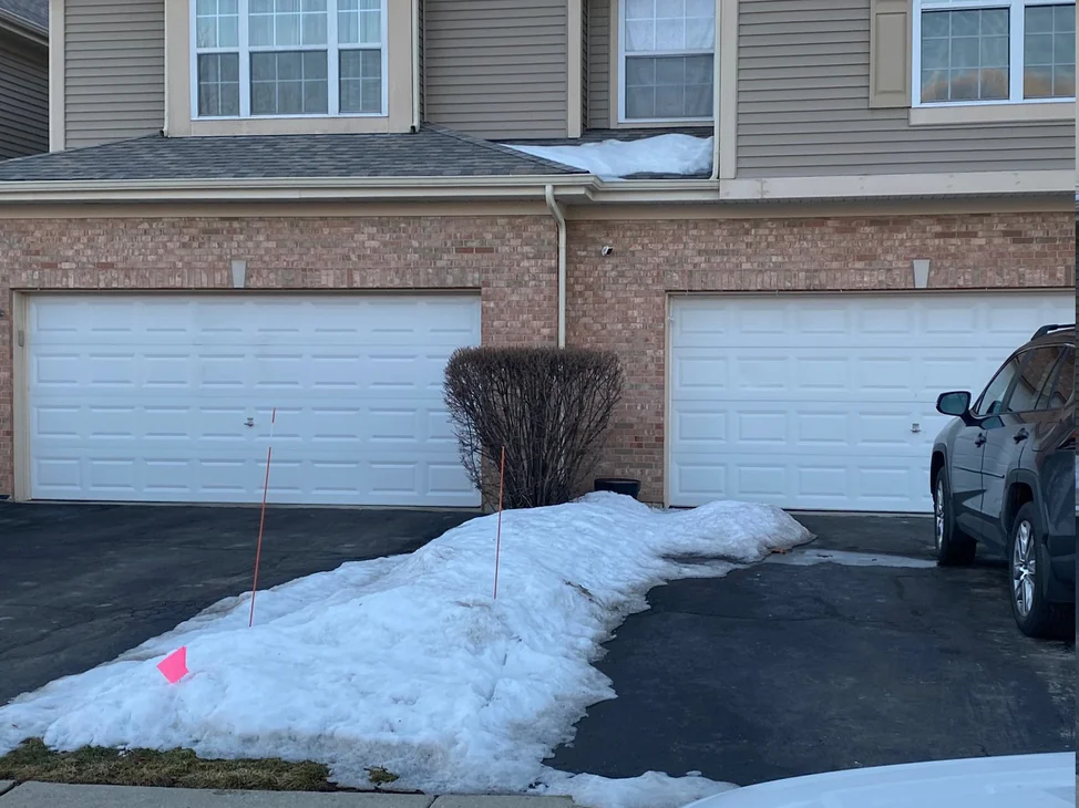 Two garage bays of the apartment row in 2019, brick running across both openings under a second storey of tan siding, snow banked along the drive
