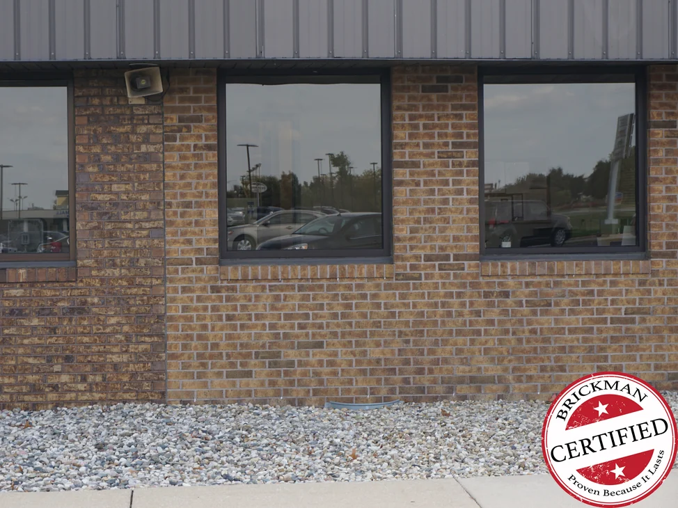 A brick elevation of the dealership in 2019, three windows set between brick piers, the wall reading as one tan and brown range from the sills down to the gravel bed