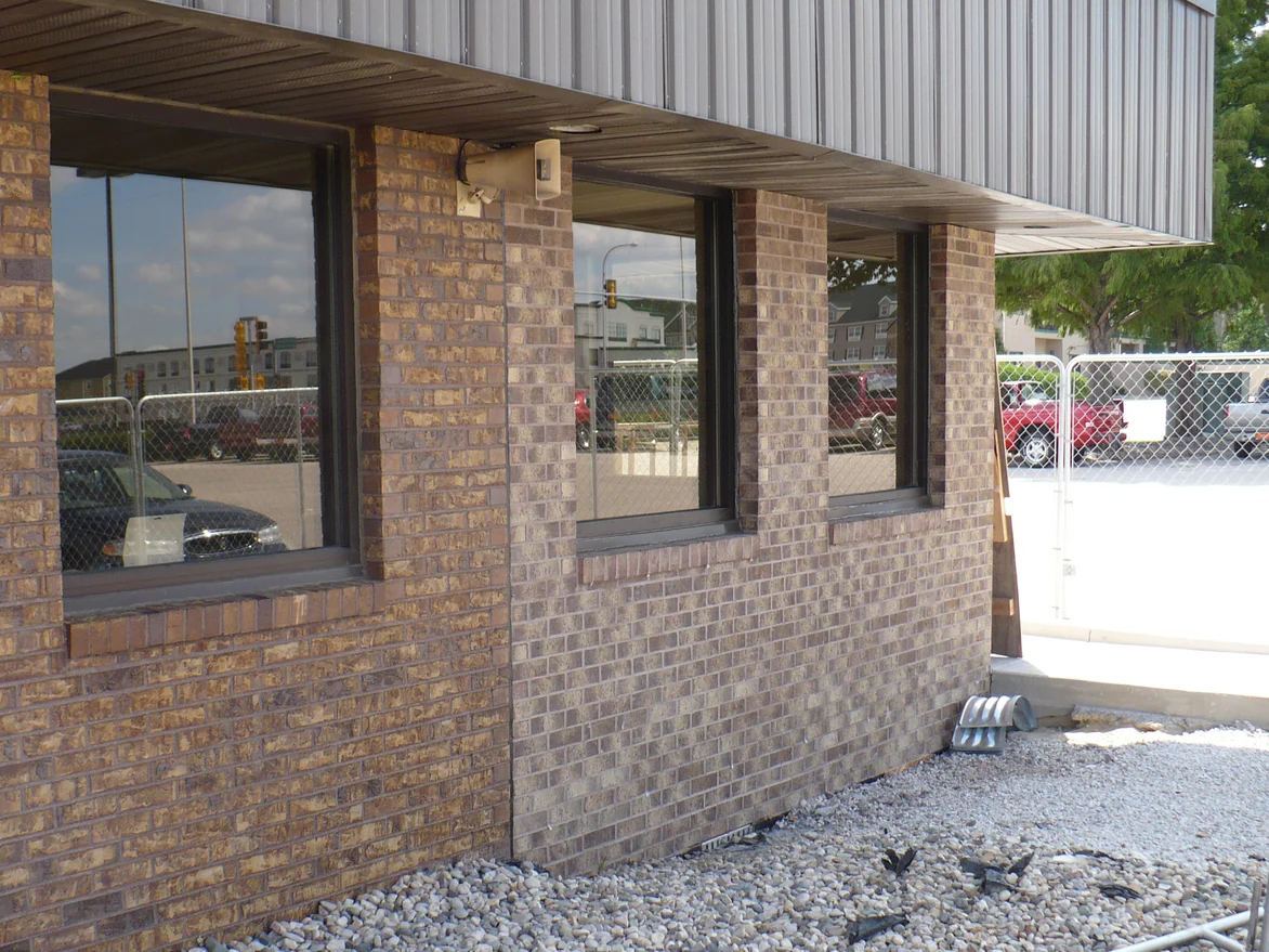 A raking view along the dealership wall before the work, the brick piers a warm tan and the wall panel beyond them a cooler grey brown