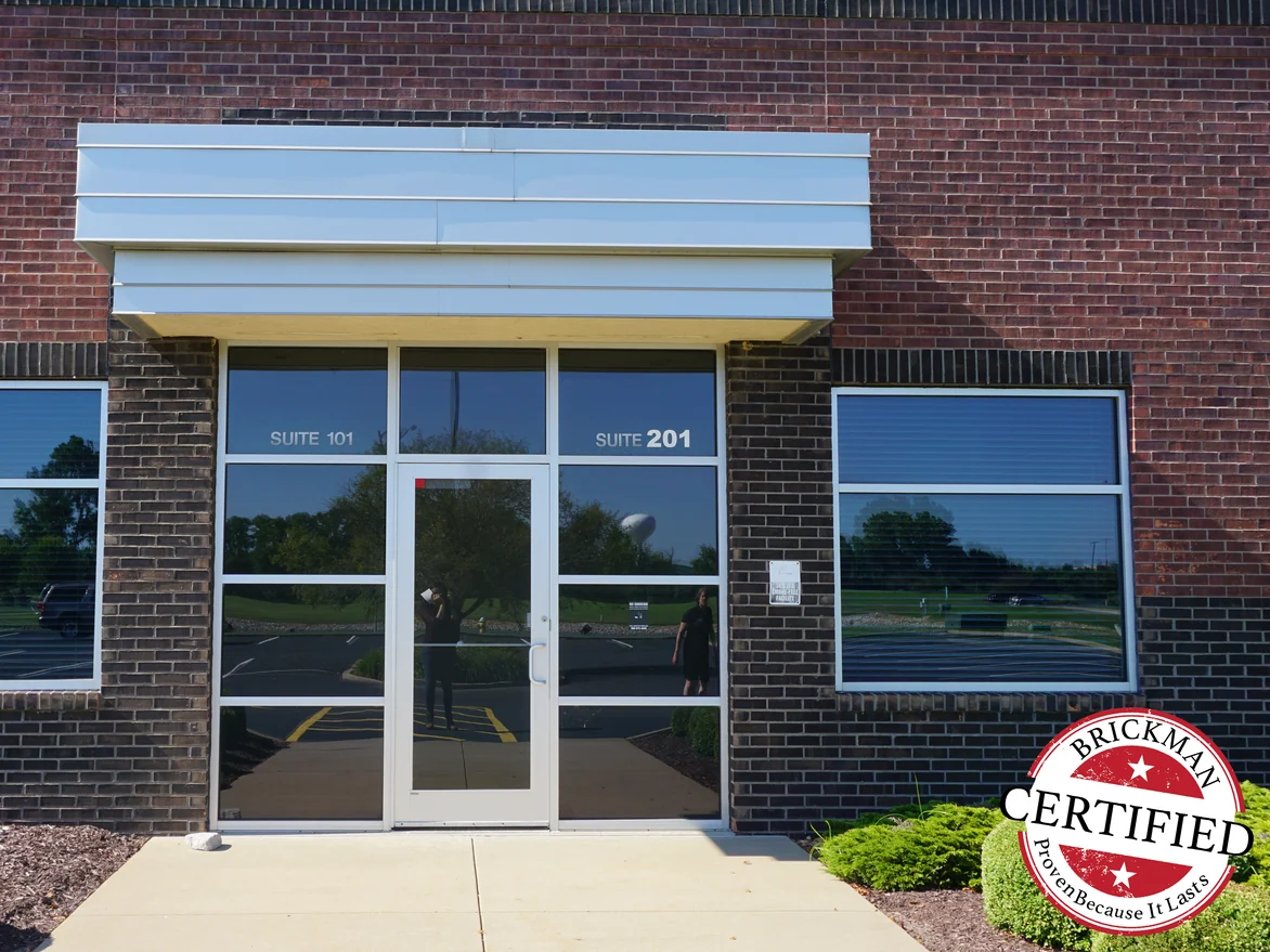 The same glazed entrance in 2019, its brick piers and base course an even dark charcoal against the red field brick, with the certification stamp at the corner