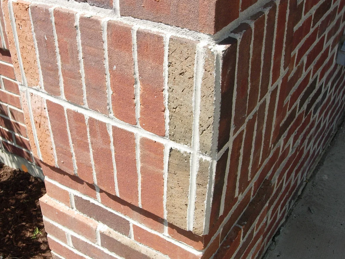 An outside corner before the work, the upright soldier course carrying one pale peach unit and one raw buff unit against red brown brick, with a mulch bed at the foot of the wall