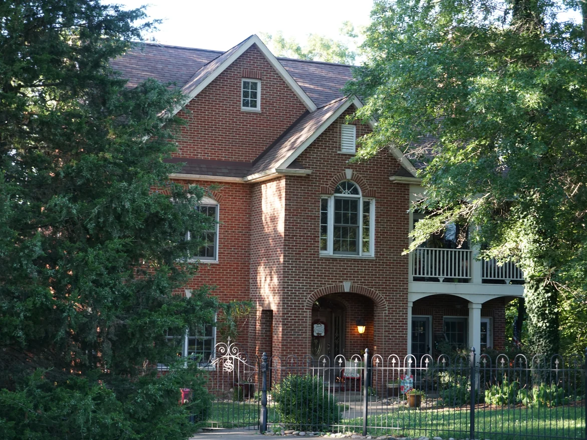 The house in 2019 seen through the trees along the front boundary, the red brick running unbroken from the gable down to the arched entry