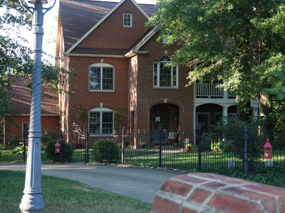 The whole front of the house from the driveway in 2019, the arched entry in the middle and a covered porch and balcony to the right, all in one red brick range