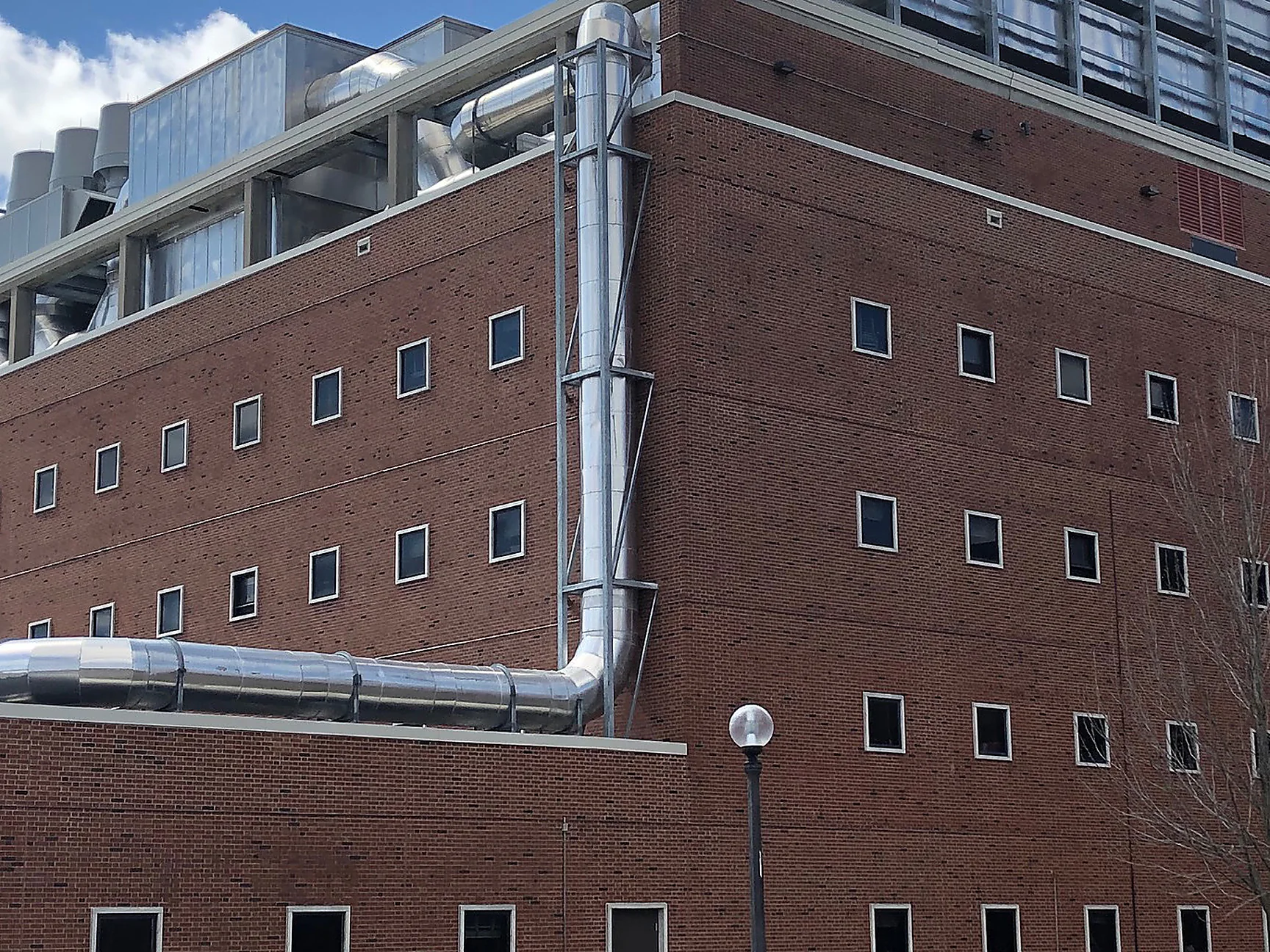 The corner of a red brick university building in 2019, five floors of small square windows under rooftop ductwork and a bank of louvred plant, a lamp post and bicycle racks at the base