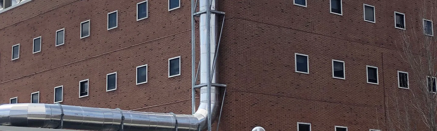 The middle floors at the corner of the red brick university building in 2019, rows of small square windows and a metal duct on the face