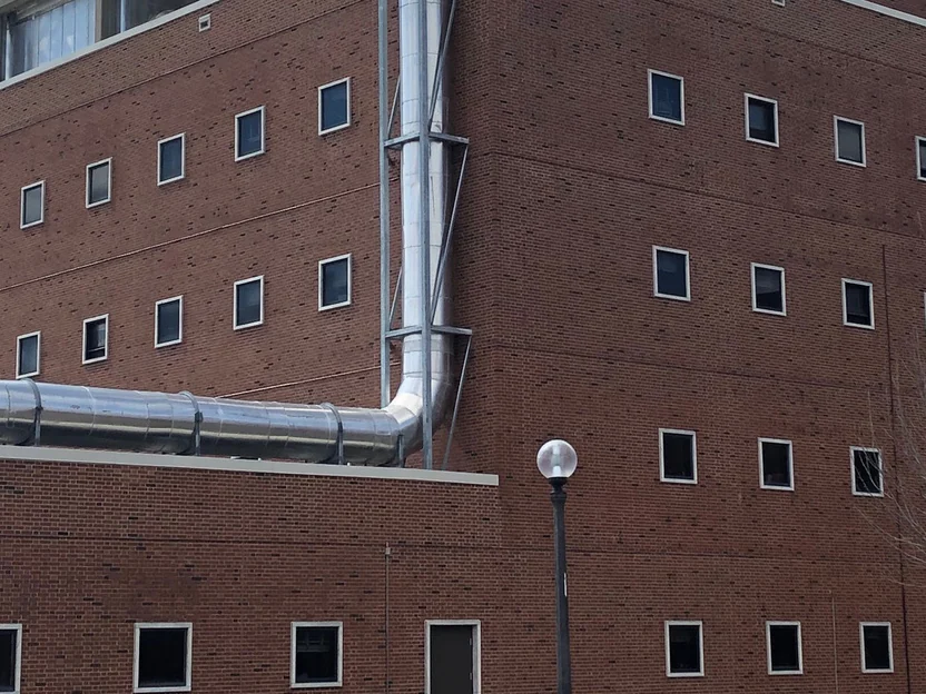 The middle floors at the corner of the building in 2019, red brick running through the corner past rows of small square windows, with a metal duct dropping down the face
