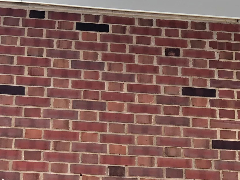 Brick at close range in 2019 under a metal parapet cap, red and brown units in every course with dark headers scattered through them