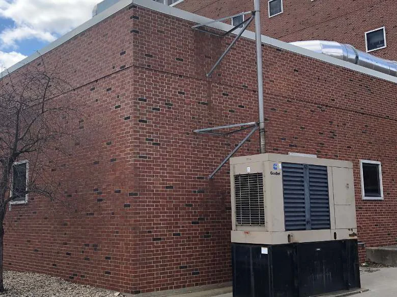 A single-story corner of the building in 2008 with a generator set against the wall, the corner brick reading lighter and flatter than the tall wing behind it