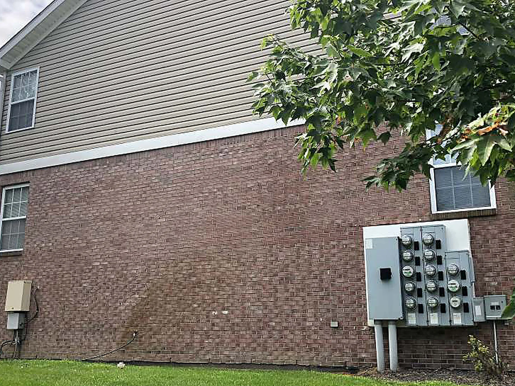 The same side elevation in 2019, the brick under the tan siding reading as one red brown range from the downspout to the meter bank, with a tree in leaf at the corner