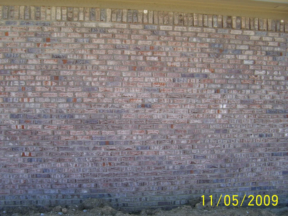 A long brick wall in 2009 under a pale soffit, its units running from cream and pink through to slate grey, with bare soil at the footing