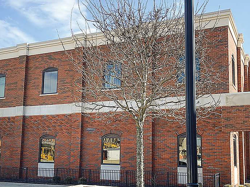 The bank in 2021, two stories of red brick with scattered dark headers carrying the length of the elevation to a canopied entrance at the far corner, a bare tree and a lamp post in front