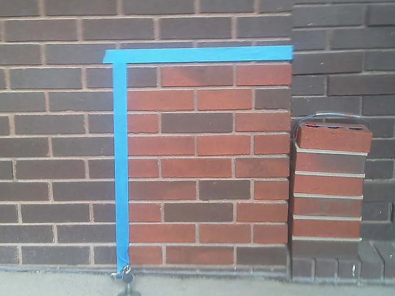 A square of the dark brown wall masked off with blue tape in 2003 and stained red, with a loose brick of the target color propped against the wall beside it
