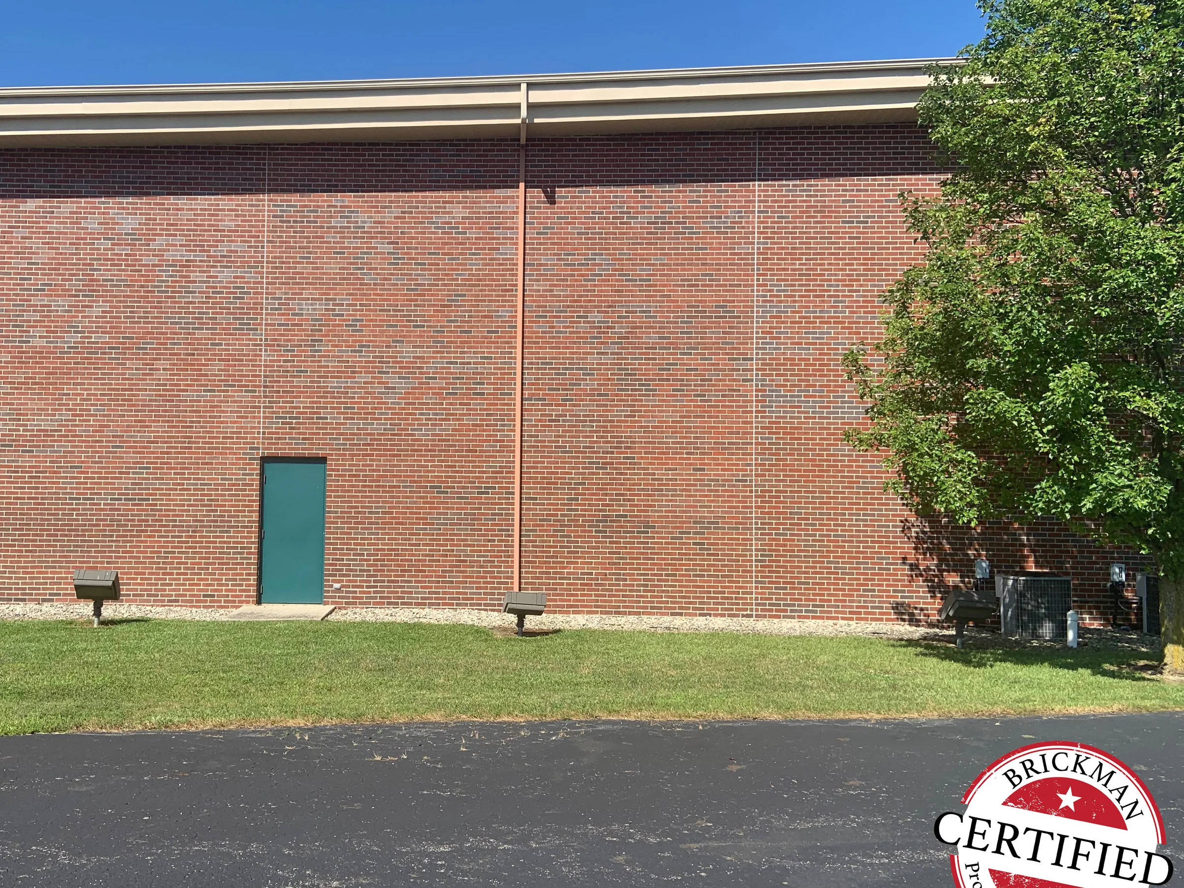 The same elevation in 2019 seen from the car park, the brick reading as one even red and grey blend from the eaves down to the gravel bed, a green service door at its foot and a mature tree at the far corner