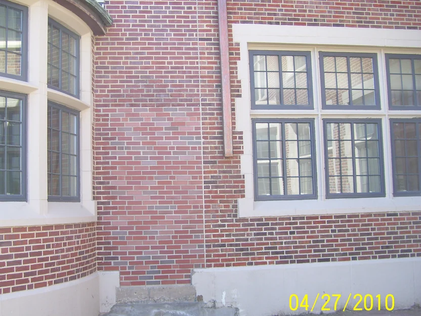 A fill-in panel of pale pink brick between a curved bay window and a copper downspout in April 2010, lighter than the red and dark brick of the original school on either side, with bare soil at the foundation