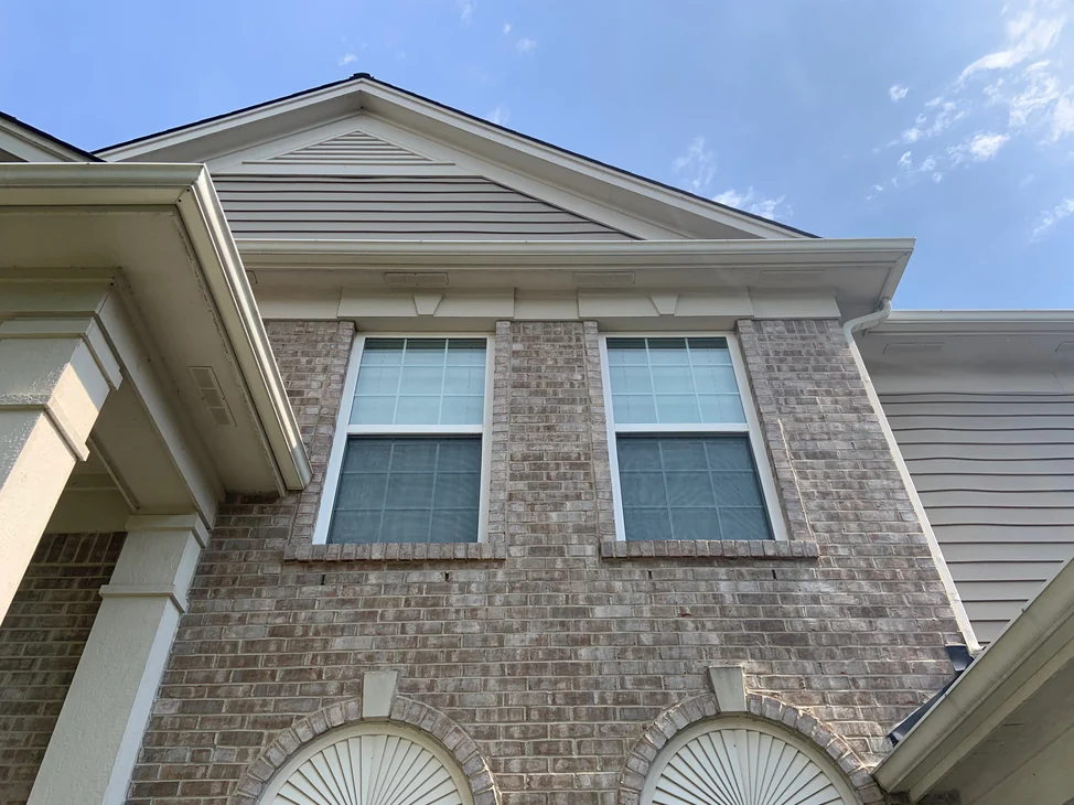 Looking up at the gable end of the house in 2019, two upper windows set in brick that carries one even tan and cream range, with siding in the gable above.