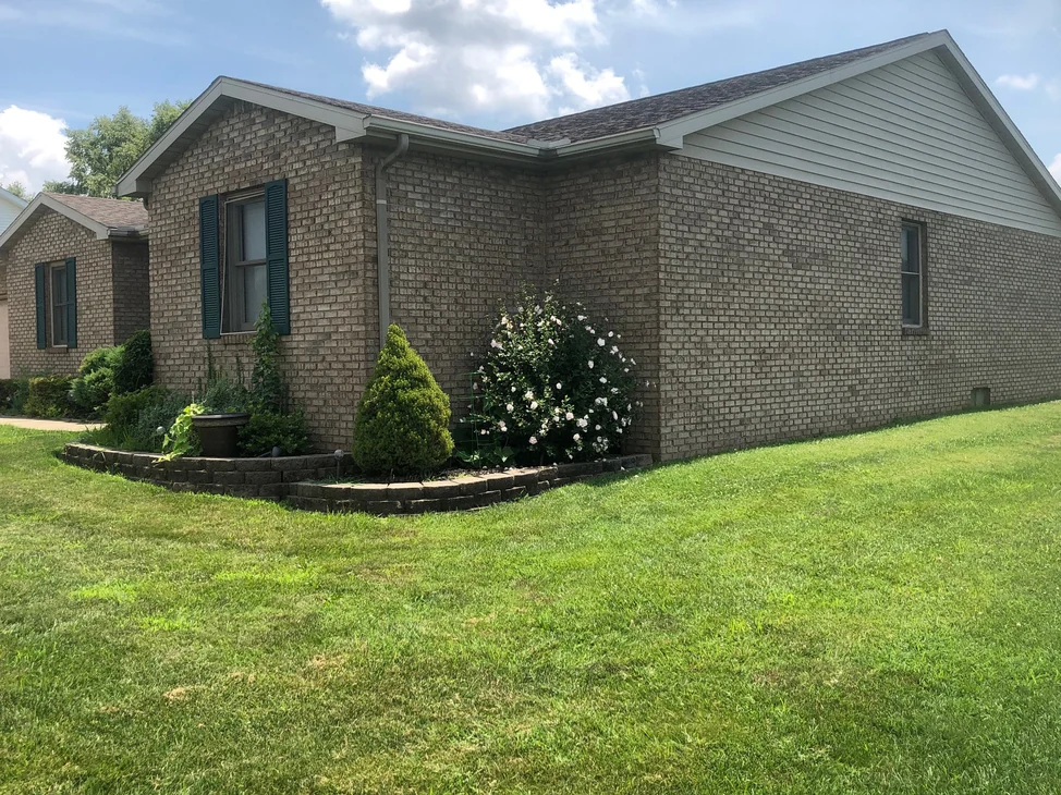 The corner of the single-story home in 2019, tan brick with a dark-shuttered window on the gable end and one long side wall running under a vinyl gable, a rose bush and a small evergreen in the bed at the corner