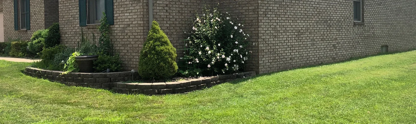 The corner of the single-story brick home in Indiana in 2019, tan brick running from a shuttered gable window along the side wall under a vinyl gable