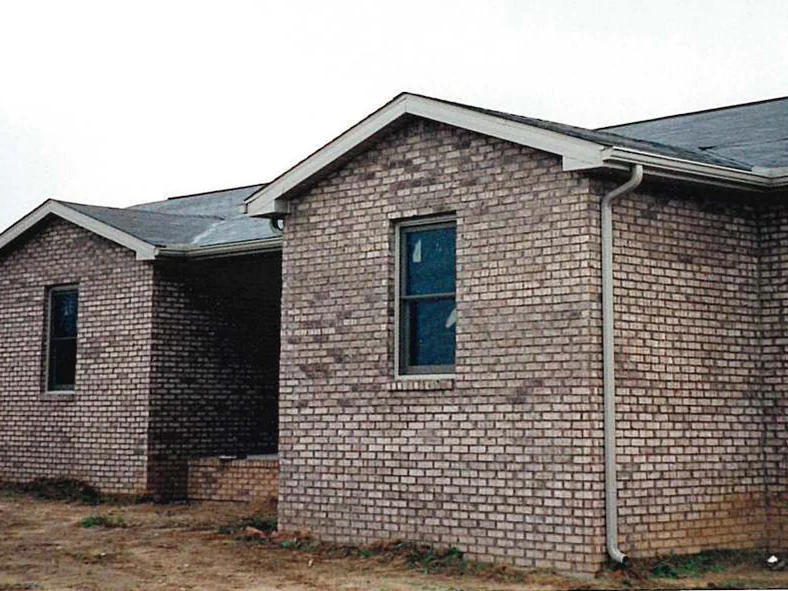 The corner of the home in December 1998 on bare graded dirt, dark units scattered through the light brick of the gable end and the side wall, a second gable beyond