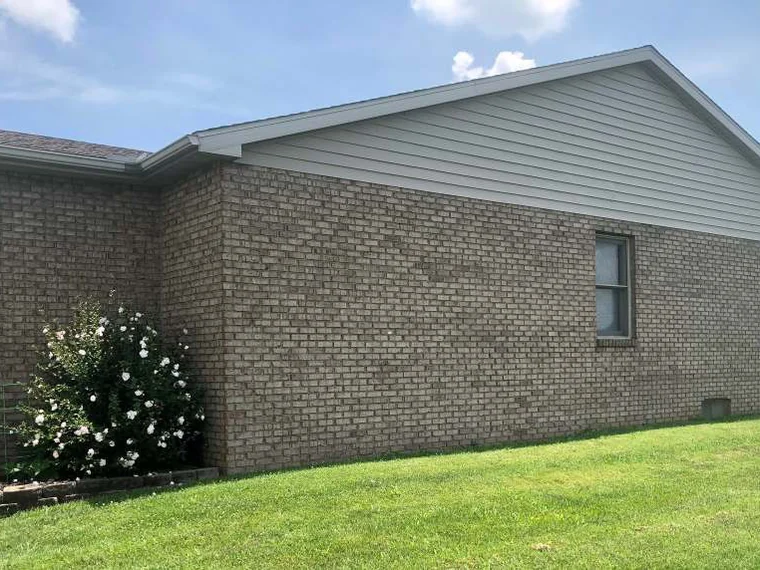The gable end of the home in 2019, one range of tan brick under a vinyl-sided gable with a single window, a white flowering shrub at the corner and lawn in front
