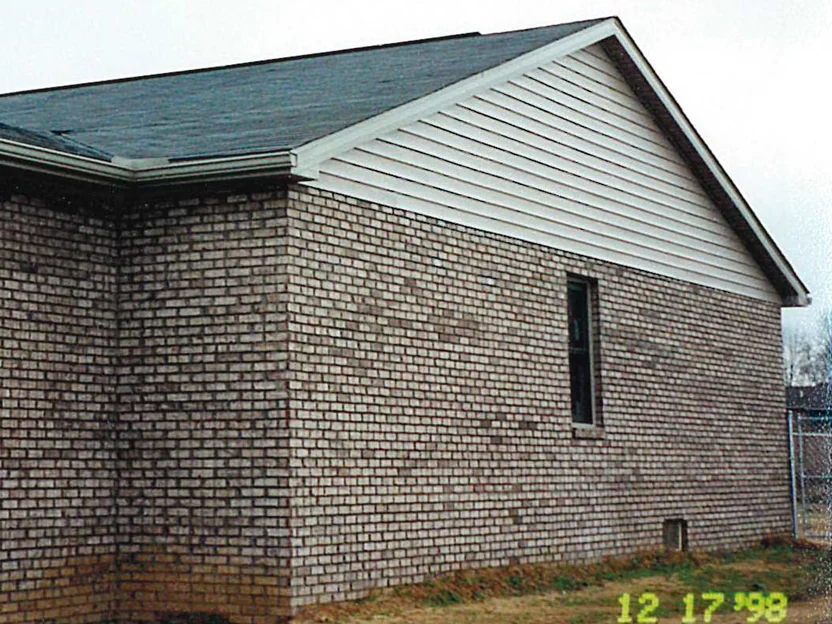 The gable end of the home in December 1998, blotchy light brick below a vinyl-sided gable with one tall window, a chain-link fence behind and the camera date 12 17 '98 in the corner