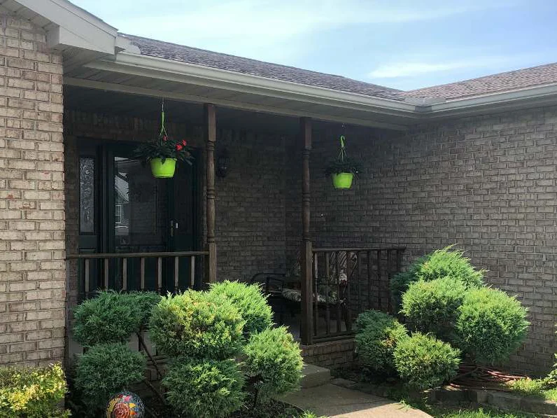 The covered front porch of the home in 2019, tan brick walls behind turned posts, hanging baskets and a row of evergreen shrubs