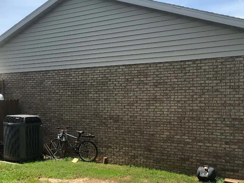 The rear wall of the home in 2019, one range of tan brick under vinyl siding, an air conditioning unit and a bicycle against the wall