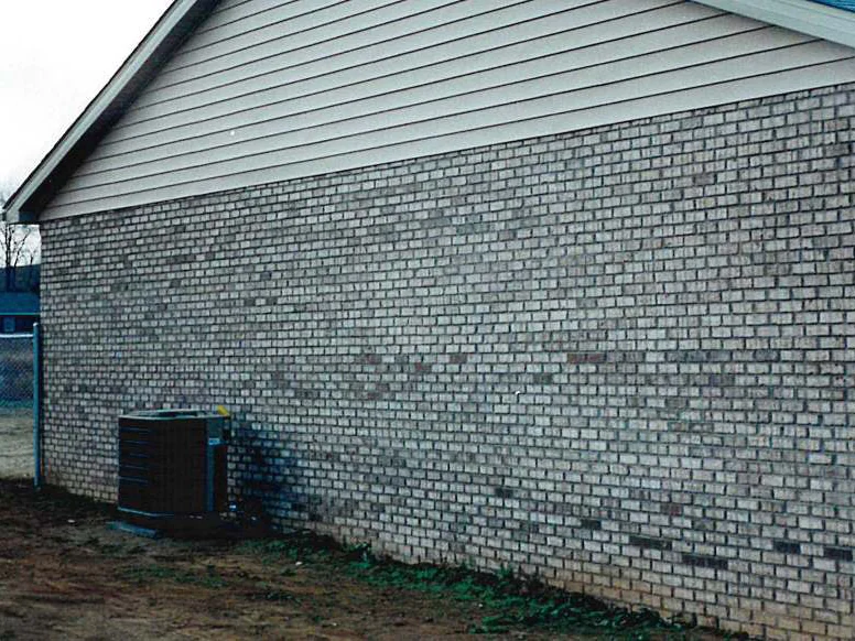 The rear wall of the home in December 1998, dark patches of brick along the base of a light blotchy wall, an air conditioning unit at the left corner