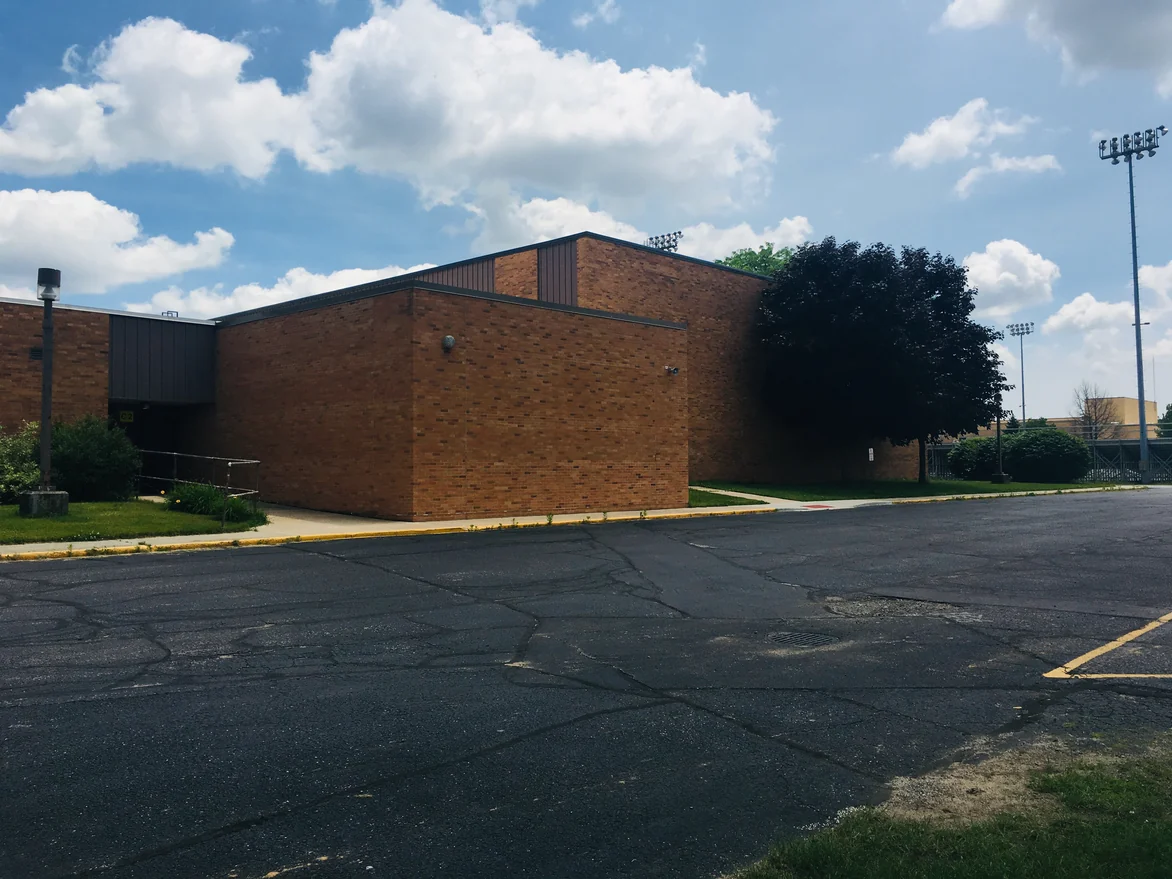 The corner of the addition seen across the parking lot in 2019, with a mature tree beside it and stadium lights standing beyond