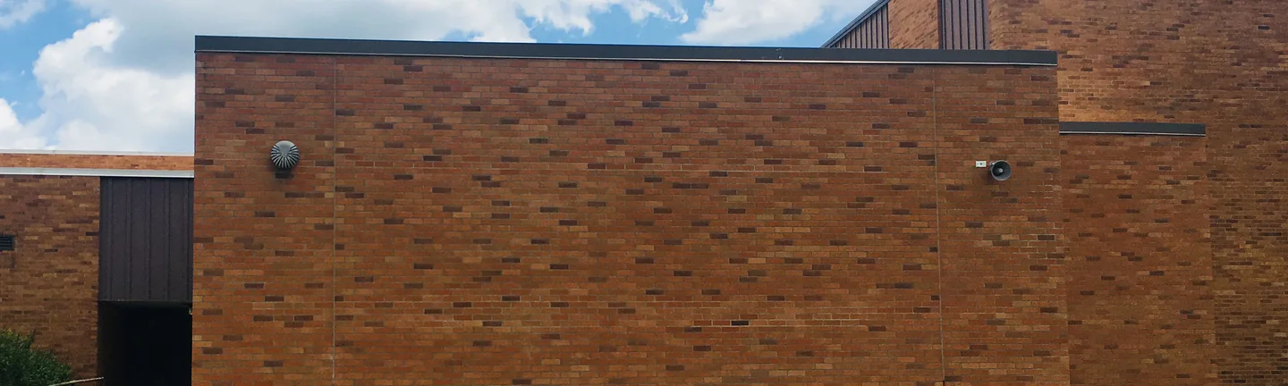 The stained wall of a school addition in 2019, dark units scattered through the brick below a flat roofline