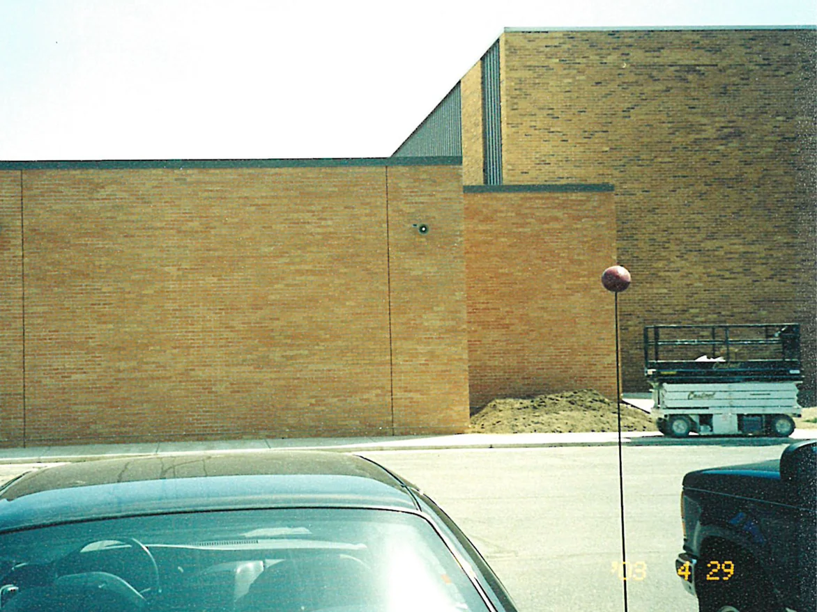 A new school wing in one flat range of brick, standing in front of a taller older block whose brick carries scattered dark units, with a scissor lift and a pile of sand at the corner